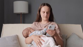 Brown haired Caucasian woman young mother holding in hands toddler daughter or son while sitting on sofa and using smartphone browsing web pages scrolling online - Powered by Shutterstock - Get 15% off with code: PIKWIZARD15
