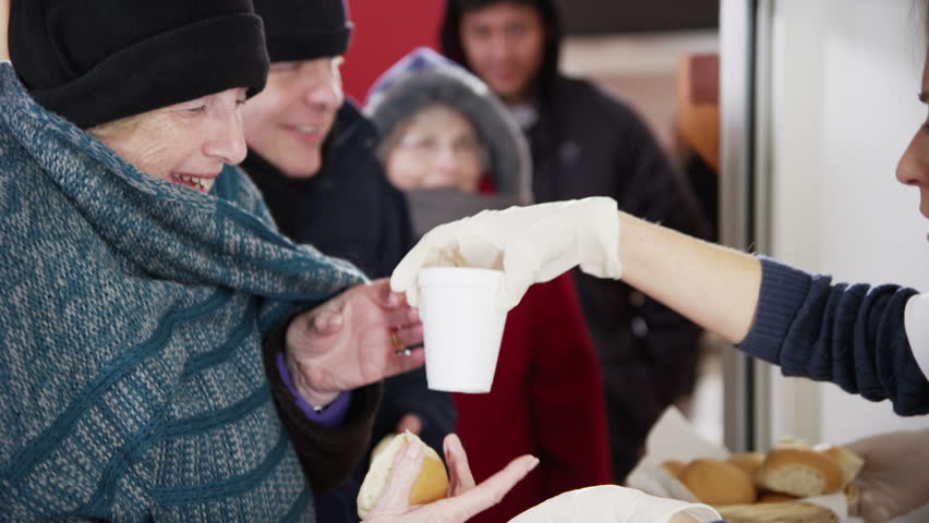 Friendly voluntary workers standing at a soup kitchen serving hatch are handing out cups of hot soup and bread to a waiting line of homeless and needy people. | Shutterstock HD Video #3583418