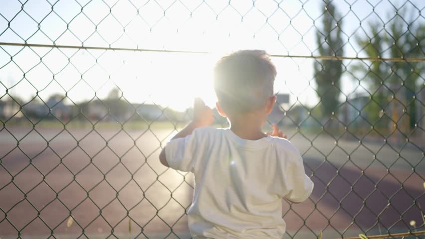boy stands near the playground. concept of happy childhood and loving family. a child holds on to the net of a sports ground, court and glare of lifestyle the sun in the background