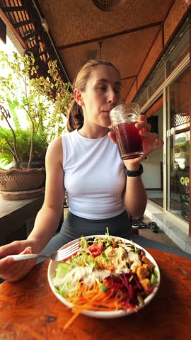 Young Hungry Pleased Woman Eating Buddha Bowl and Drink Kombucha in Restaurant. Smiling Girl Eating Fast Food and Enjoying. Thailand, Pai