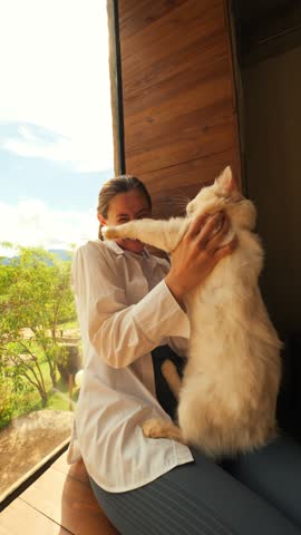 Young Beautiful Woman Sits on the Window of a Countryside House, Hugs a Fluffy White Cat and Smiling in the Sunlight