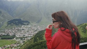 Young, attractive brunette, wearing sunglasses, enjoys a panoramic view from the summit of a mountain to a small town located in the heart of a mountainous valley. Georgia. Caucasus Mountains. Slow - Powered by Shutterstock - Get 15% off with code: PIKWIZARD15