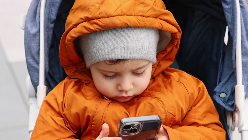 baby boy kid sitting in trolley using smartphone. mobile phone addiction concept, new generation.child in autumn jacket with hat and hood on head.focused infant outside, in park, walking time.