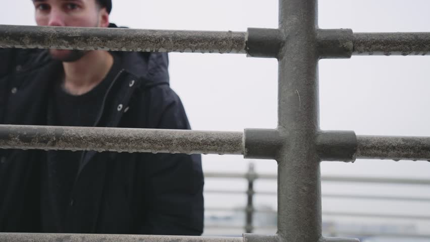 Man looking at camera through frozen metal fence in winter day outdoors. Portrait of handsome bearded man in warm clothes standing on city street alone, exhaling stream of air from mouth, stalker
