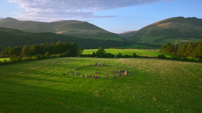People visiting ancient stone circle at golden hour with mountain background. Castlerigg, Lake District National Park, Cumbria, UK
