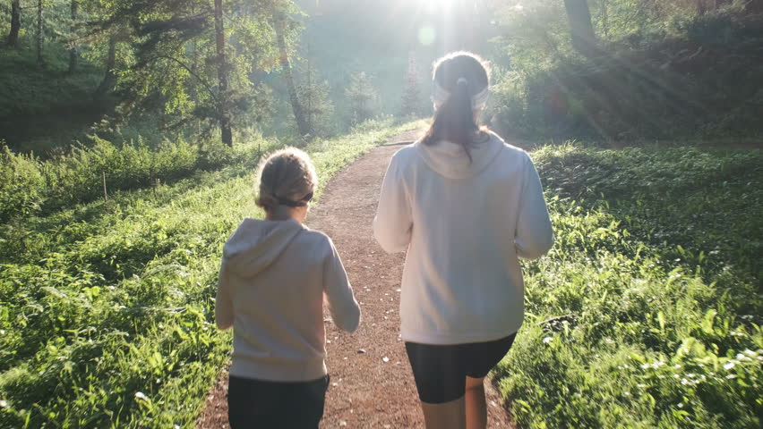 Rear view of mother and her young daughter joyfully run together along a sunlit path in the park ar dawn, enjoying surrounding nature and happy time together, slow motion. Healthy lifestyle concept