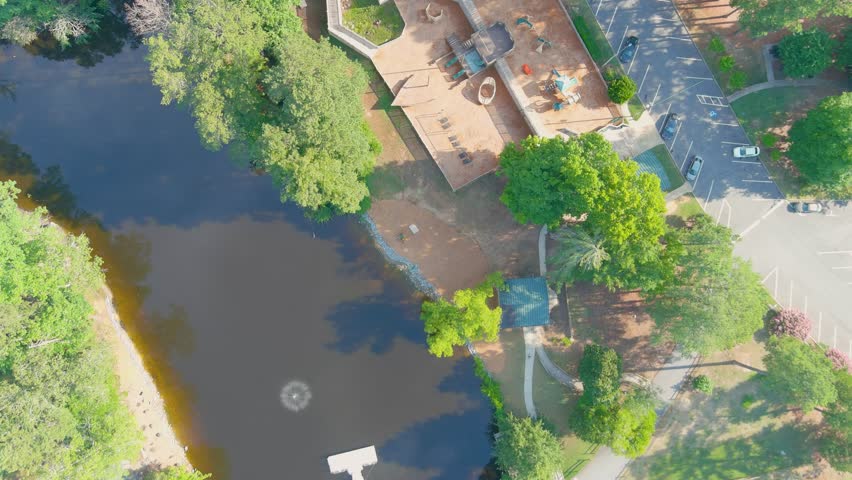 stunning aerial footage of a gorgeous summer landscape in the park with a lake and a water fountain and a brown wooden bridge surrounded by pink trees and lush green trees and grass with blue sky
