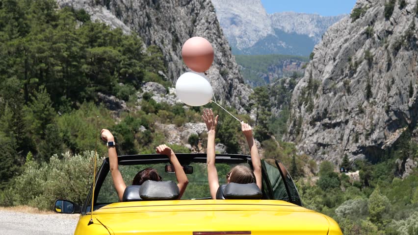 Two young woman with hands up enjoying vacation driving picturesque mountain view road