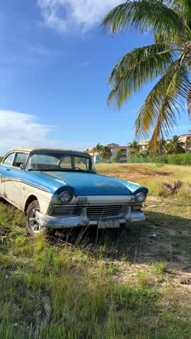 VARADERO, CUBA - DECEMBER 2, 2024: An old American car standing under a palm tree, illuminated by the sun. A retro car. Blue classic American car on the beach. 4К