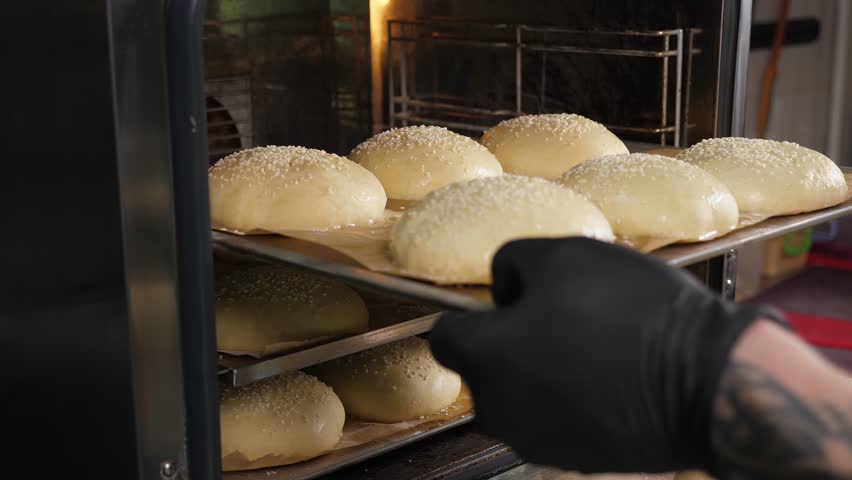A close-up of the chef puts a baking tray with burger buns in the oven in the kitchen of the restaurant. The process of cooking burgers in a restaurant. Baking sesame buns in the oven.