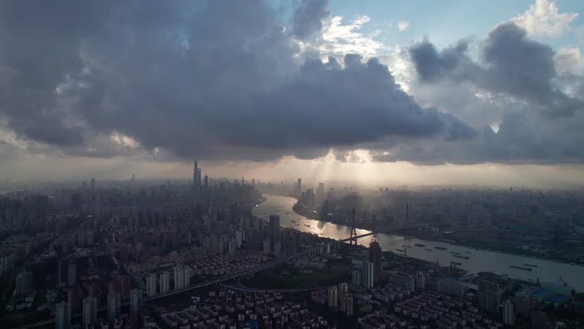 Drone view of Shanghai city in sunny cloudy rainy day at sunset, high angle view of ships sailing on Huangpu river and modern buildings with golden lights moving on sky, 4k time lapse footage.