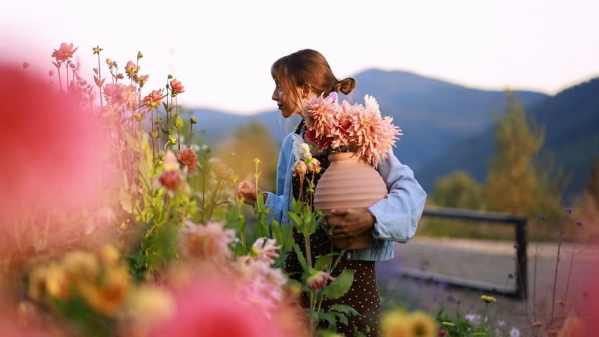 A woman gracefully collects vibrant blooms in a serene mountain garden, holding a clay vase filled with freshly picked flowers. The surrounding greenery and soft mountain backdrop