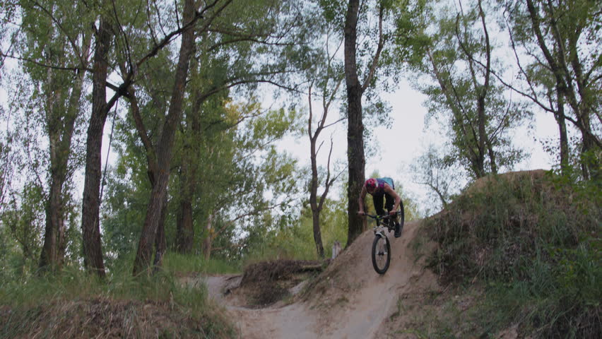 Courageous human with shirt fluttering in wind riding up hill at speed and jumping over hole in free flight. Cross-country athlete performing professional level stunt in wild nature conditions.