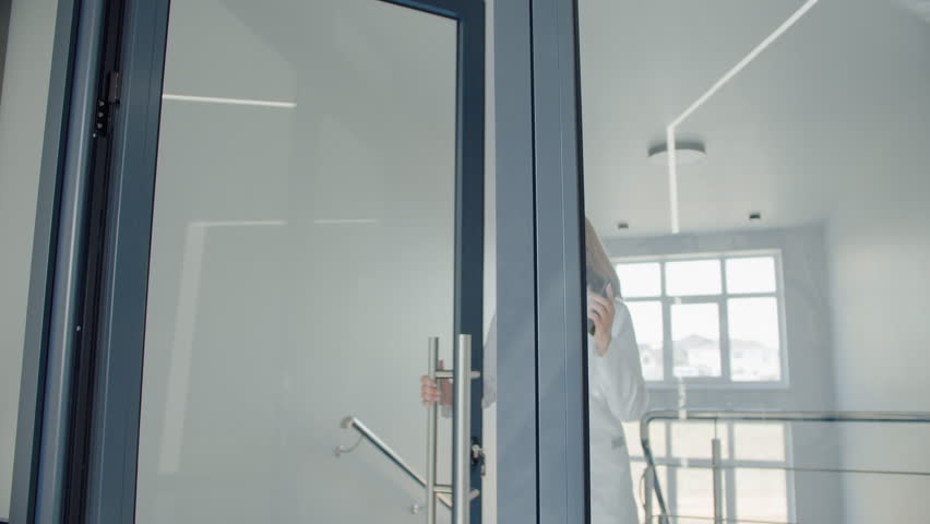 Experienced hospital employee opening door from staircase to hall while getting called to operating room. Cardiologist moving quickly through clinic with white interior while holding phone by ear.