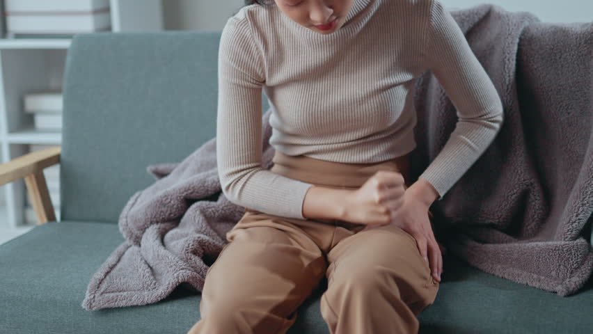 Young woman sitting on a sofa massages her leg, seeking relief from intense pain. Discomfort radiates through her body as she struggles with the symptoms of leg pain