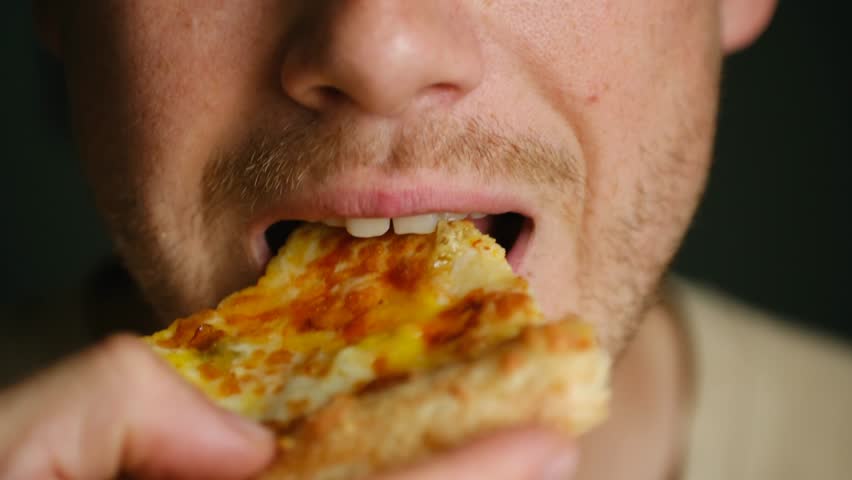 Bearded man bites off slice of cheesy pizza resting at home. Guy chews enjoying delicious taste of unhealthy snack with crispy edges for dinner