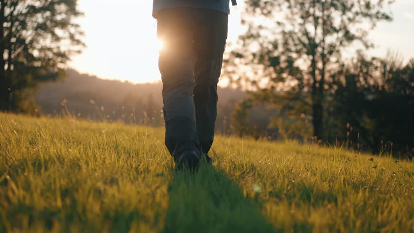 Rear back view steady shot male feet stepping on grassy meadow hill overlooking mountains in sunrise or sunset light. Man wearing hiking boots walks on clover grass. Male hiker explorer, sunny morning