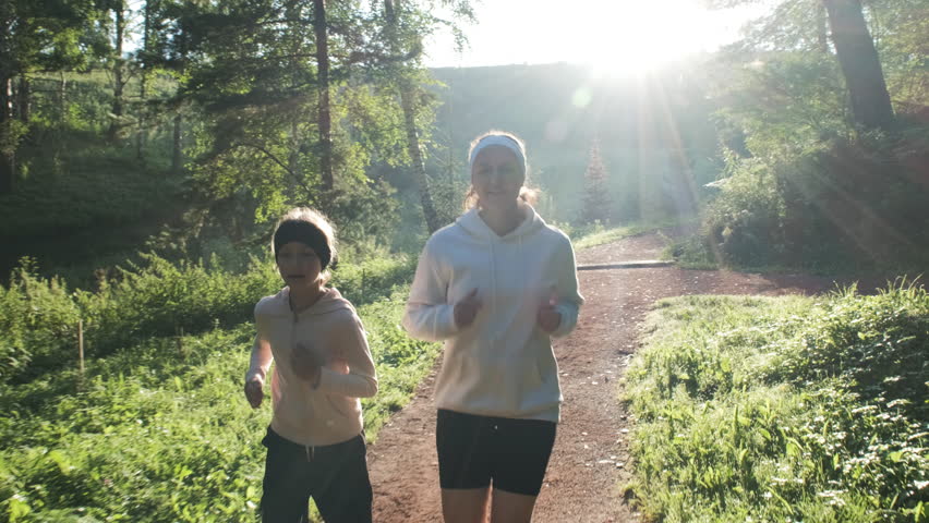 Woman and her young daughter happily jogging on a sunlit path at dawn, embracing nature and precious moments together, slow motion. Healthy lifestyle concept