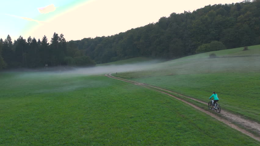 AERIAL: Woman biking through a foggy countryside with a dog running alongside her. Lady and her doggo pass the gentle mists that roll across green fields by the forest on a beautiful autumn morning.