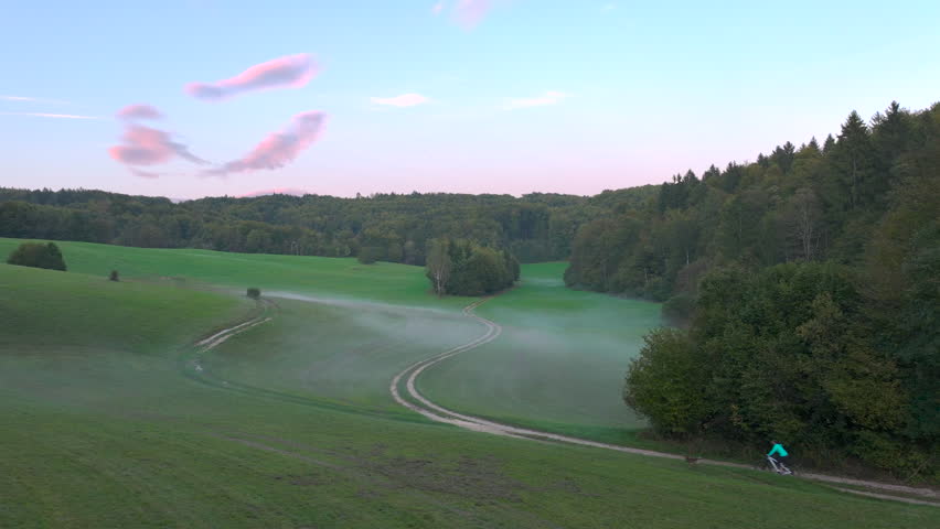 AERIAL: Riding bike accompanied by dog at dawn across a misty field by forest. A young woman during an early workout with her faithful brown doggo by her side in a beautiful misty autumn morning.
