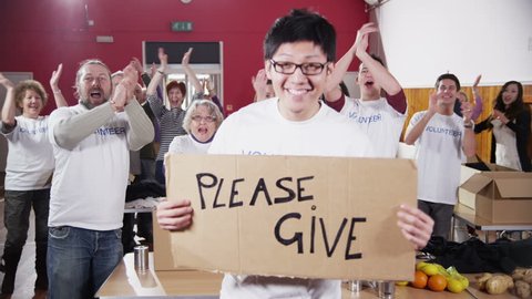 A male charity volunteer of Asian ethnicity holds up a "Please Give" sign and smiles at the camera as his fellow volunteers applaud and cheer in the background. In slow motion.