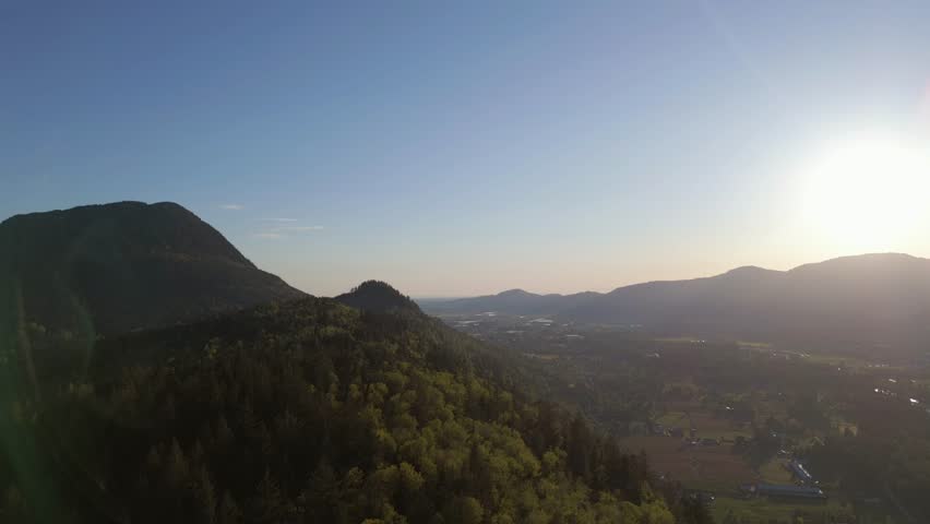 Aerial view of mountain valley with lush green trees. Farms, houses, and a town are nestled in the valley below. The sun is setting behind the mountains creating a warm glow. British Columbia, Canada.