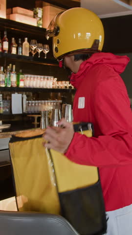 Vertical shot of Asian deliveryman in uniform taking food bag in chic restaurant for transportation
