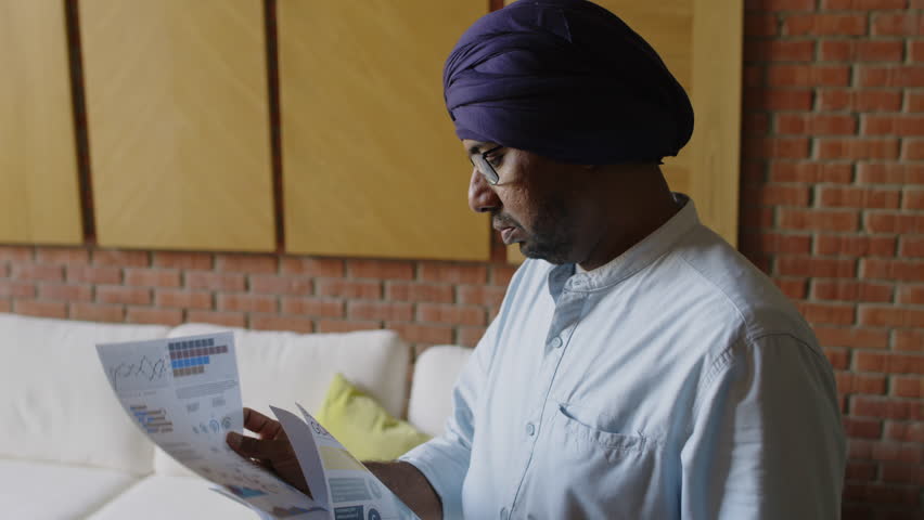 Tilt down view of Indian boss wearing turban comparing papers with analysis at work in modern office