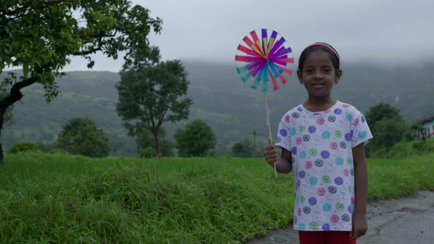 Bhandardara, Maharastra, India- August 11th- 2024-Compact high speed shot of Indian young girl holding a paper windmill in hand and smiling towards the camera.with  mountainous background.