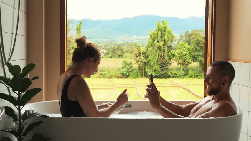 Romantic Bath in Hotel: Couple in Bathtub, Boyfriend Taking Photos as Girlfriend Blows Bubbles, Overlooking Scenic Rice Fields in Thailand, Capturing Playful Honeymoon Moments