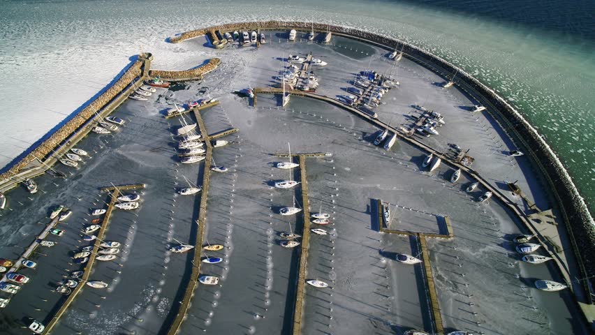 Drone circles around over boat marina in Denmark, in wintertime. The marina is packed with ice, and there is also packed with ice right outside the harbour. It is a sunny winter day