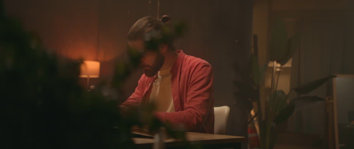 Footage young Caucasian man sitting at desk in evening and using digital tablet. Male graphic designer working in living room. Freelance work. Remote job. Camera moving around
