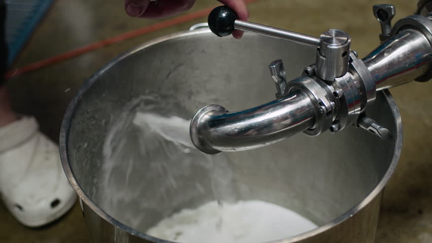 Closeup of stainless steel container being filled with liquid from pipe. Unrecognizable worker operating valve to control filling process