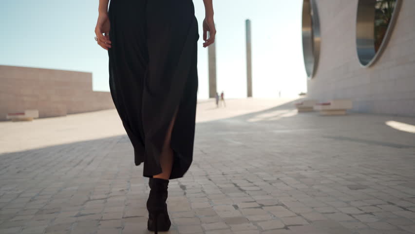 Sensuous woman in black dress gesturing towards camera and dancing on street during sunny day . Beautiful young female model is displaying graceful freestyle moves outdoors. 