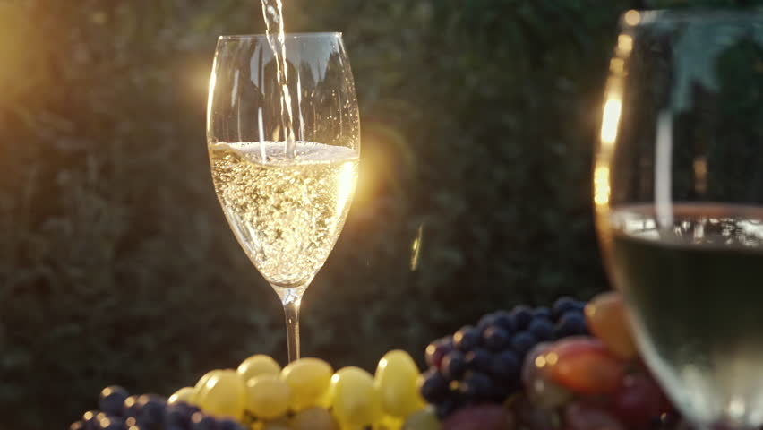 Stunning view of pouring white wine into glass goblet with fresh grape bunch background, sun light breaks through vineyard. 