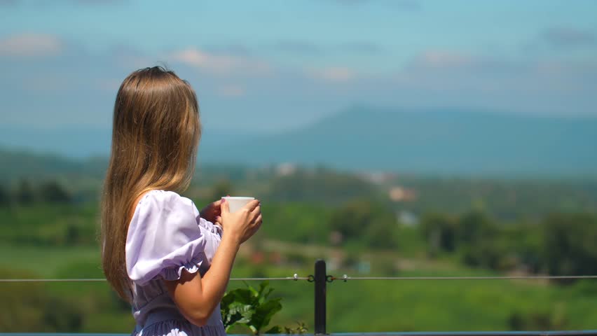 Young woman enjoying peaceful morning with cup of coffee while standing on balcony, with serene mountain landscape and greenery in background.
