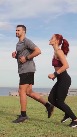 Vertical. Caucasian couple running on the beach wearing sportswear. A sporty man and a healthy woman enjoy exercising on the shoreline. Energetic friends having a cardio workout jogging at coast line