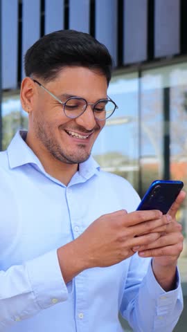 Vertical. Hispanic young entrepreneur or business man smiling and using a cell phone app, wearing executive shirt and glasses. latin professional male browsing on internet with smartphone at workspace