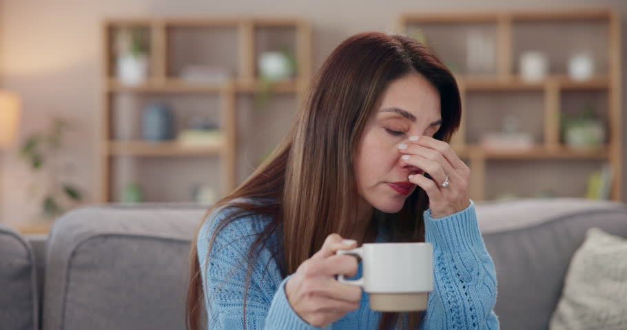Woman, home and headache with coffee on sofa with stress, unhappy and pain in living room. Female person, couch and migraine in lounge with overthinking, burnout and tired or frustrated with tea