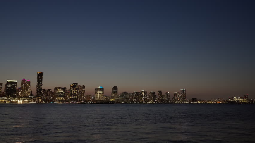 A wide view of the New Jersey skyline at dusk, with city lights reflecting on the calm waters of the Hudson River. The sky transitions from twilight to night.