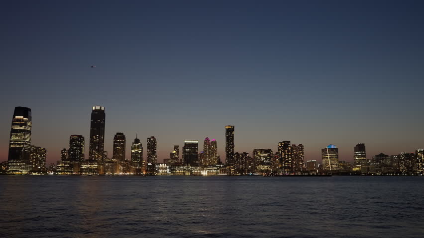 The New Jersey skyline illuminated at night, reflecting on the Hudson River, under a twilight sky. The city lights create a vibrant urban scene with a serene water view.