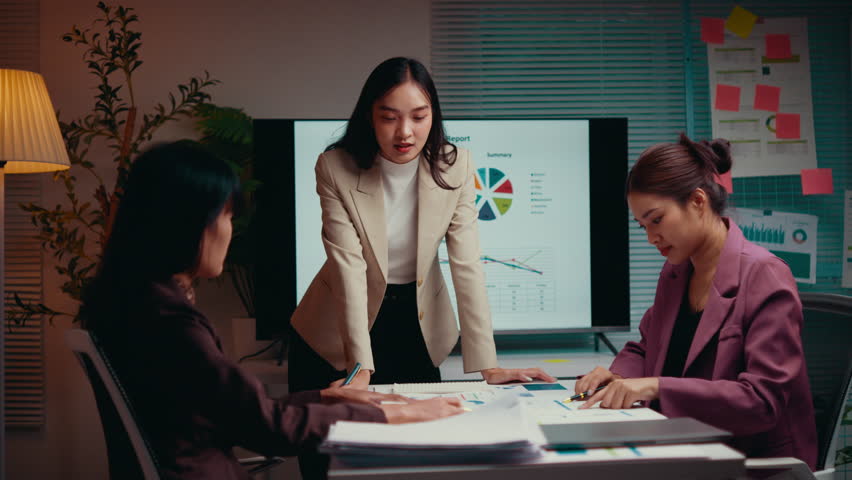 Businesswoman standing and gesturing while explaining a project to two colleagues during a night meeting in the office, fostering teamwork and collaboration through discussion and analysis