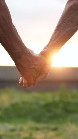 Elderly couple enjoys romantic moment as they stand hand in hand, silhouetted against the warm glow of the setting sun. Close up holding hands. Helping hand. Care old person. Vertical video