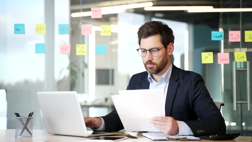 Confident businessman working checks documents using a laptop computer in modern business office. Busy serious financier in formal suit is doing paperwork while sitting at the desk at the workplace