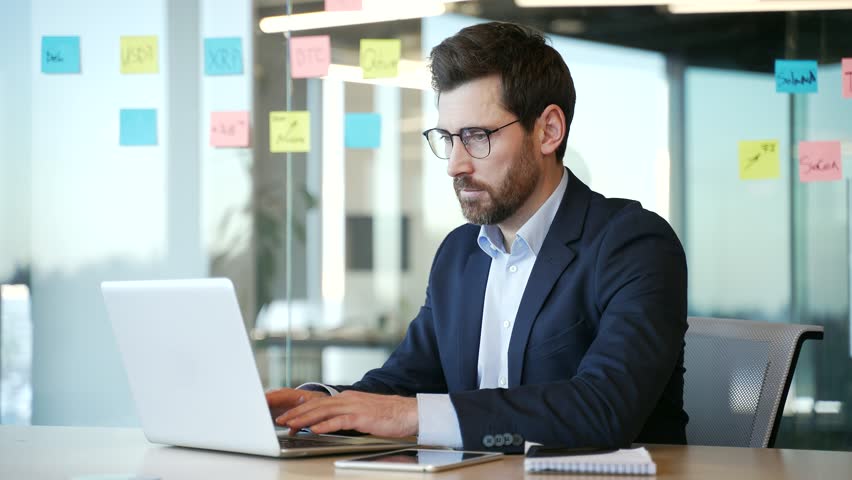 Overworked businessman in a formal suit suffering from eye strain and headache working on laptop sitting at workplace in business office. Tired man takes off glasses and massages, rubs bridge of nose