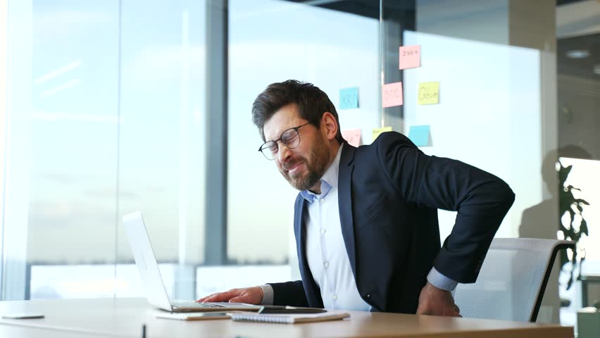 Tired businessman in a formal suit suffering from back pain while working on laptop sitting at desk at workplace in business office. Upset worker massages painful lower back muscles. He is exhausted