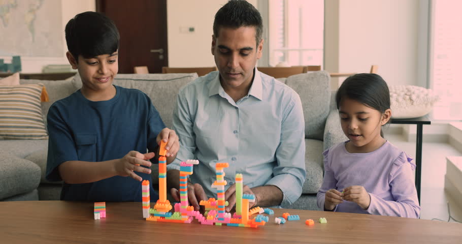 Focused Indian dad and two kids constructing city from toy blocks, building towers in home living room, talking to preteen son and little daughter girl, smiling, laughing
