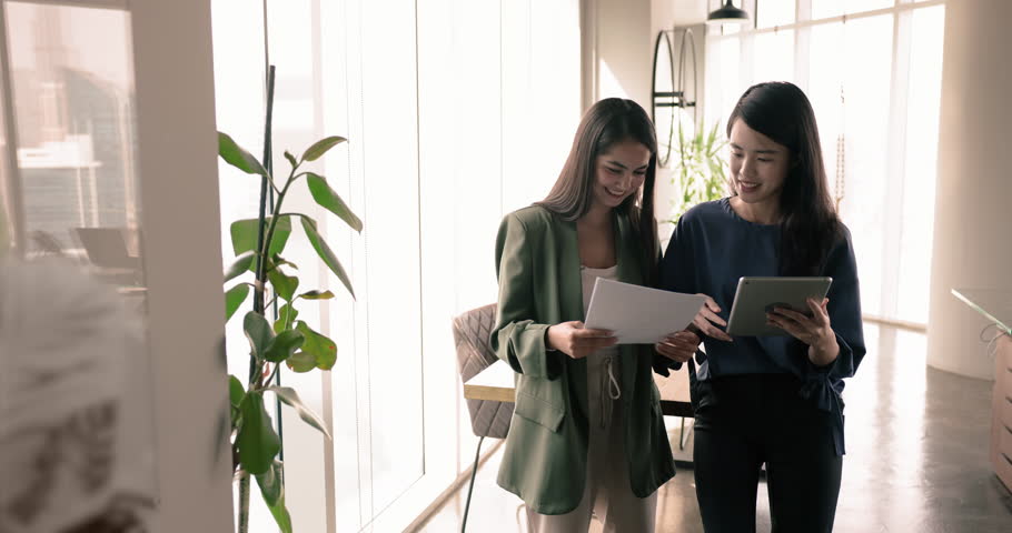 Two multiethnic staff members Asian and Hispanic women colleagues lead talk met in modern office holding paper with sales results and tablet discuss joint project, share strategy and opinion. Teamwork