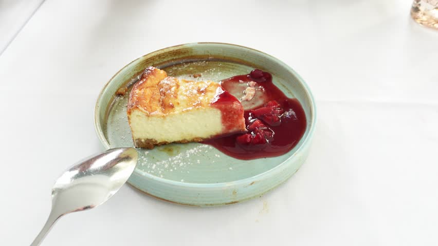 Detail of a cheesecake with raspberry jam on a green plate on the white tablecloth of a restaurant table. A hand with a metal spoon cuts off a piece to eat it