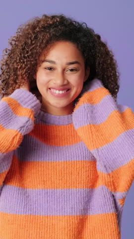Vertical video, Smiling young woman with curly brown hair wearing striped cozy sweater adjusting her hair standing on lilac background in studio